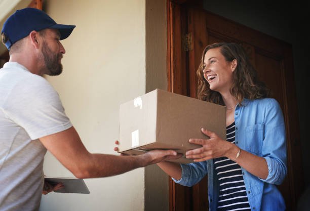 A male delivery driver in a baseball cap hands a cardboard package to a smiling woman standing at her doorstep.