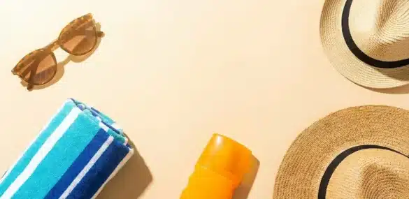 Flat lay view of summer essentials including tortoise-shell sunglasses, a striped blue towel, sunscreen, and two straw hats on a beige background.